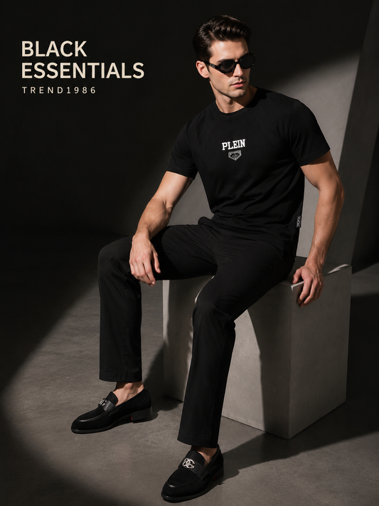 Seated male model wearing a black logo T-shirt, sunglasses and leather shoes in a dark gallery setting